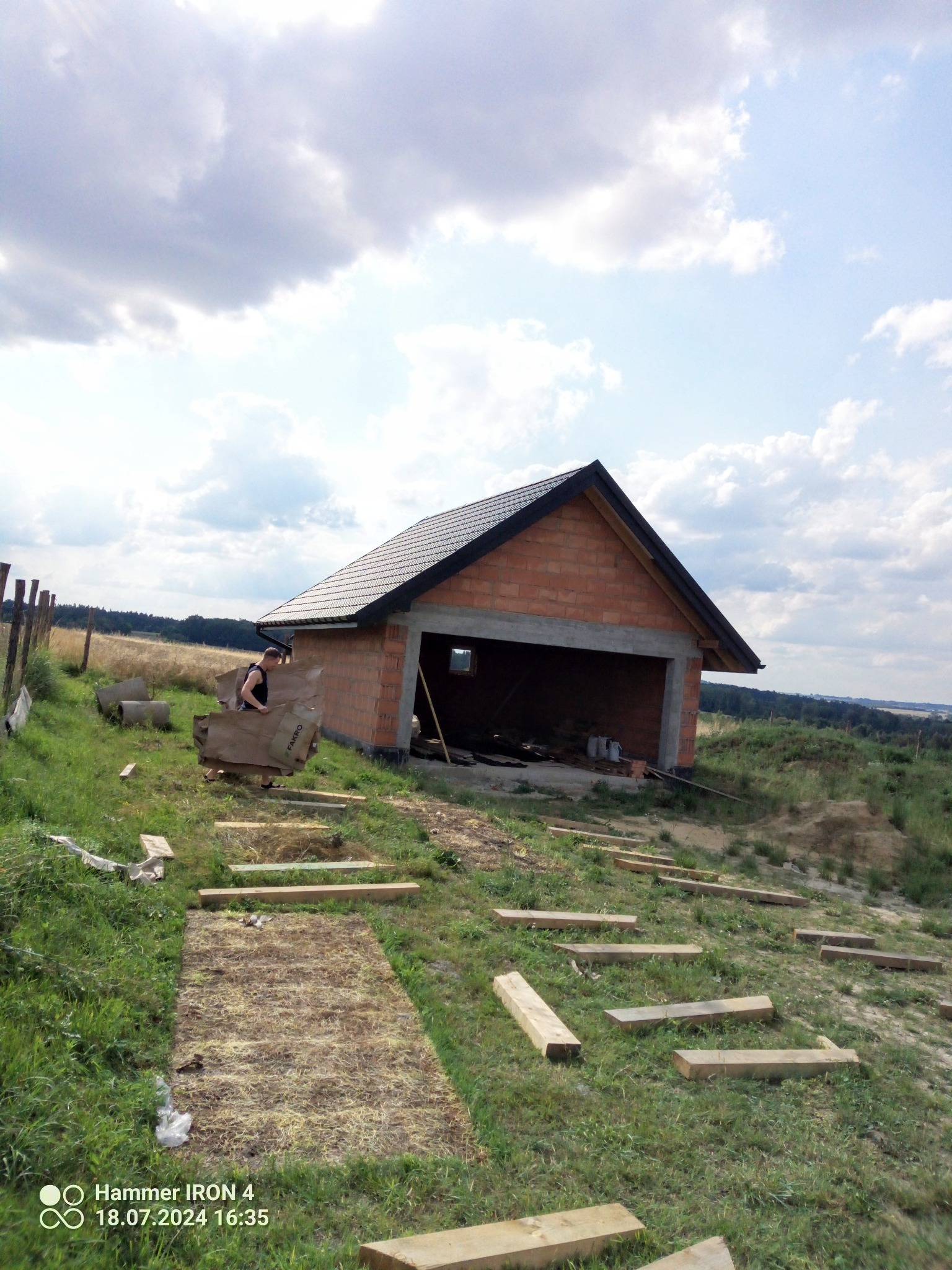 Budowa domu murowanego w Zagajach w trakcie realizacji, widoczne ściany z czerwonej cegły i czarny dach, pracownik przenosi materiały budowlane.
