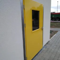 Żółte drzwi zewnętrzne z małym okienkiem, zamontowane w białej ścianie budynku z widoczną kratką odpływową wzdłuż ściany i kostką brukową na chodniku.