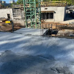 Świeżo wylany beton na budowie, widoczny dźwig, szalunki i zbrojenia, w tle budynki mieszkalne.