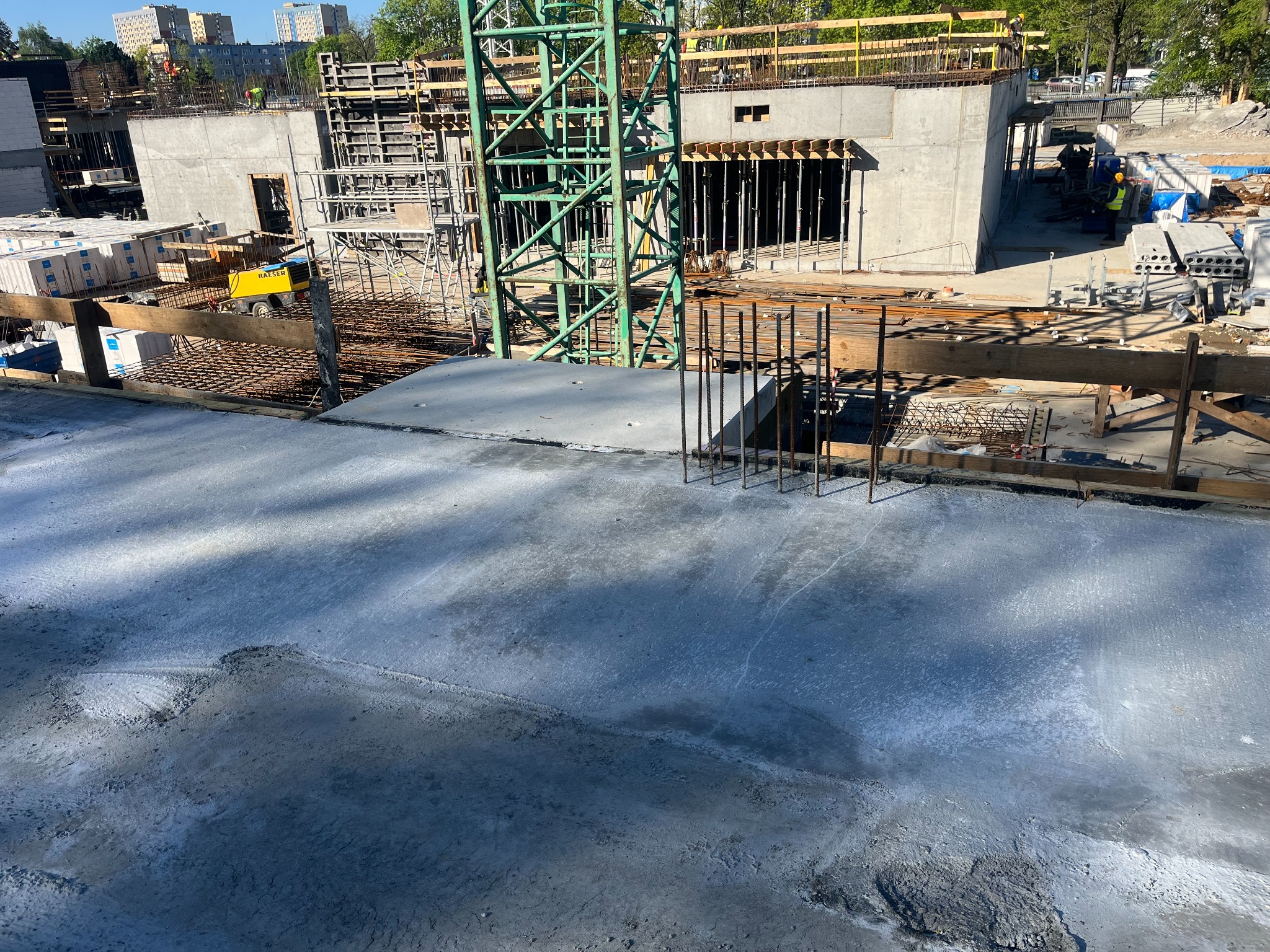 Świeżo wylany beton na budowie, widoczny dźwig, szalunki i zbrojenia, w tle budynki mieszkalne.