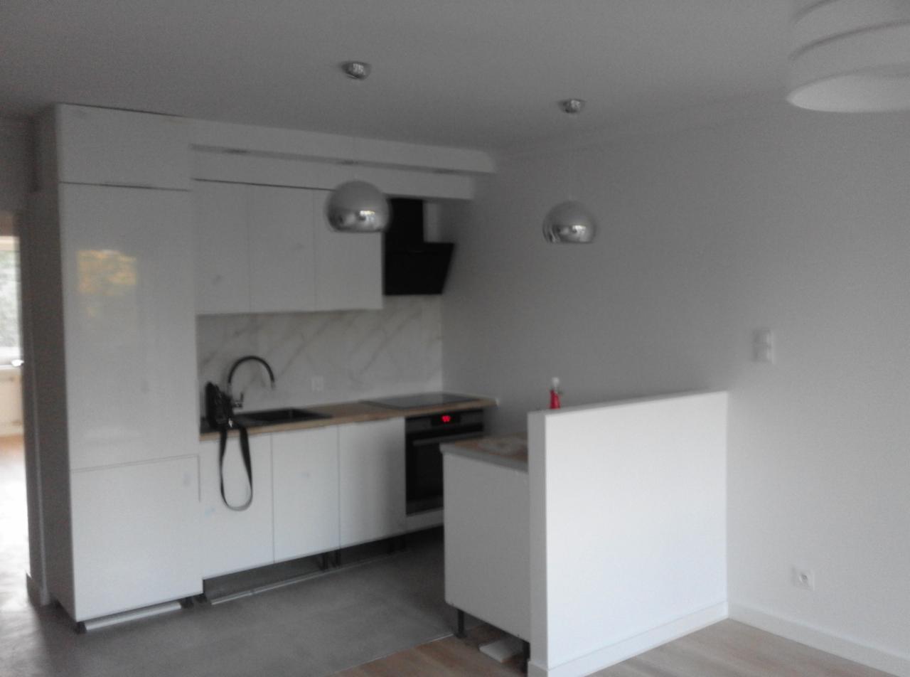 Nowoczesna, minimalistyczna kuchnia z białymi szafkami, marmurowym backsplashiem i srebrnymi lampami wiszącymi. Widoczny czarny zlew z baterią i płyta indukcyjna. Świeżo wyremontowane wnętrze...