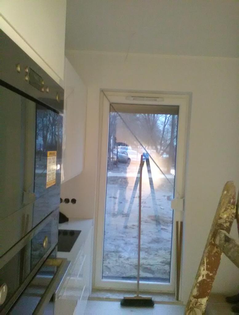 Wnętrze w trakcie prac wykończeniowych: widoczna drabina oparta o ścianę przy oknie, obok stoi miotła, a w tle zabudowa kuchenna z połyskiem.