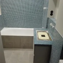 Łazienka w trakcie remontu: ściany i obudowa umywalki wyłożone mozaiką w odcieniach błękitu, biała wanna, szare płytki podłogowe, blat pod umywalkę z otworem wypełnionym zaprawą.