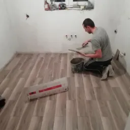 Glazurnik układa podłogowe płytki imitujące drewno w łazience, widoczny kubeł z klejem i paczka płytek, przygotowane do montażu.