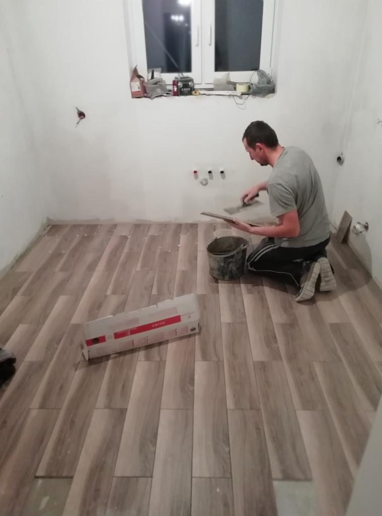 Glazurnik układa podłogowe płytki imitujące drewno w łazience, widoczny kubeł z klejem i paczka płytek, przygotowane do montażu.