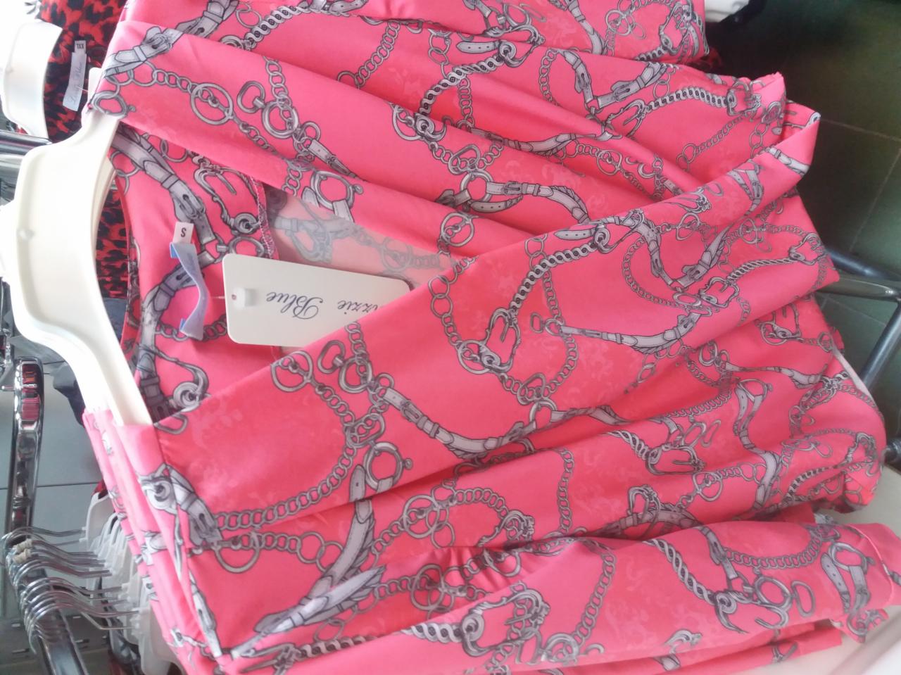 Stos ubrań w kolorze fuksji z nadrukiem łańcuchów, zawieszonych na wieszaku w sklepie odzieżowym, widoczna metka z nazwą marki.