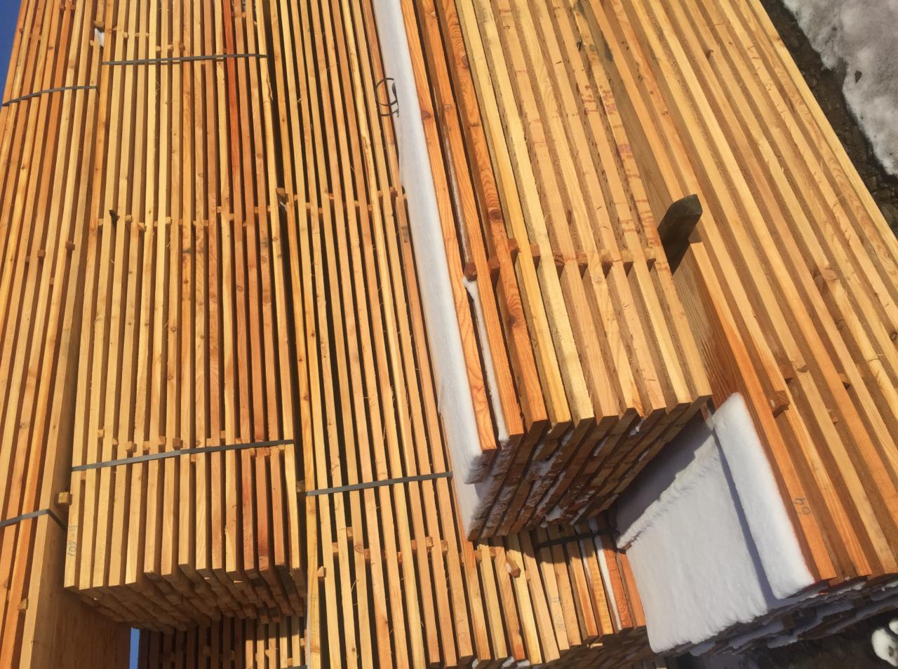 Stosy świeżo przetartego drewna budowlanego, częściowo przykryte śniegiem, zabezpieczone taśmami. Widoczne słoje i naturalna kolorystyka drewna.