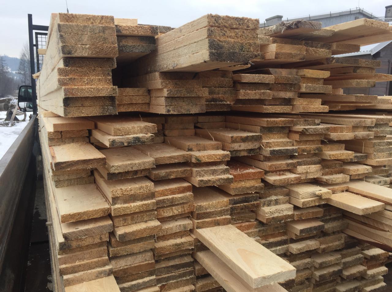 Stos świeżo przetartego drewna budowlanego w skrzyni samochodu dostawczego, widoczne ślady po cięciu i fragmenty kory, w tle zabudowania i drzewa.