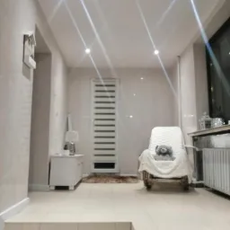 Jasne wnętrze łazienki z beżowymi płytkami, podwieszanym sufitem z punktowym oświetleniem, białymi meblami i dekoracyjnym fotelem z pluszowym misiem.