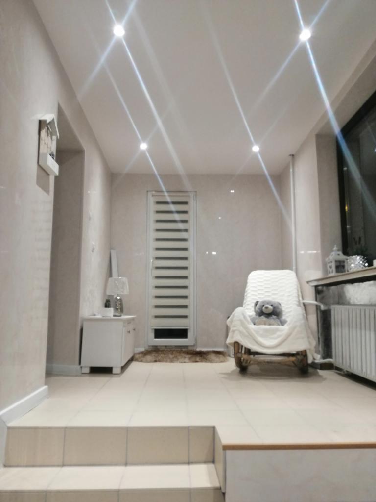 Jasne wnętrze łazienki z beżowymi płytkami, podwieszanym sufitem z punktowym oświetleniem, białymi meblami i dekoracyjnym fotelem z pluszowym misiem.
