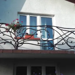 Kuta balustrada balkonowa z motywem drzewa i liści, z doniczką z czerwonymi kwiatami, na tle białej elewacji domu z oknami.