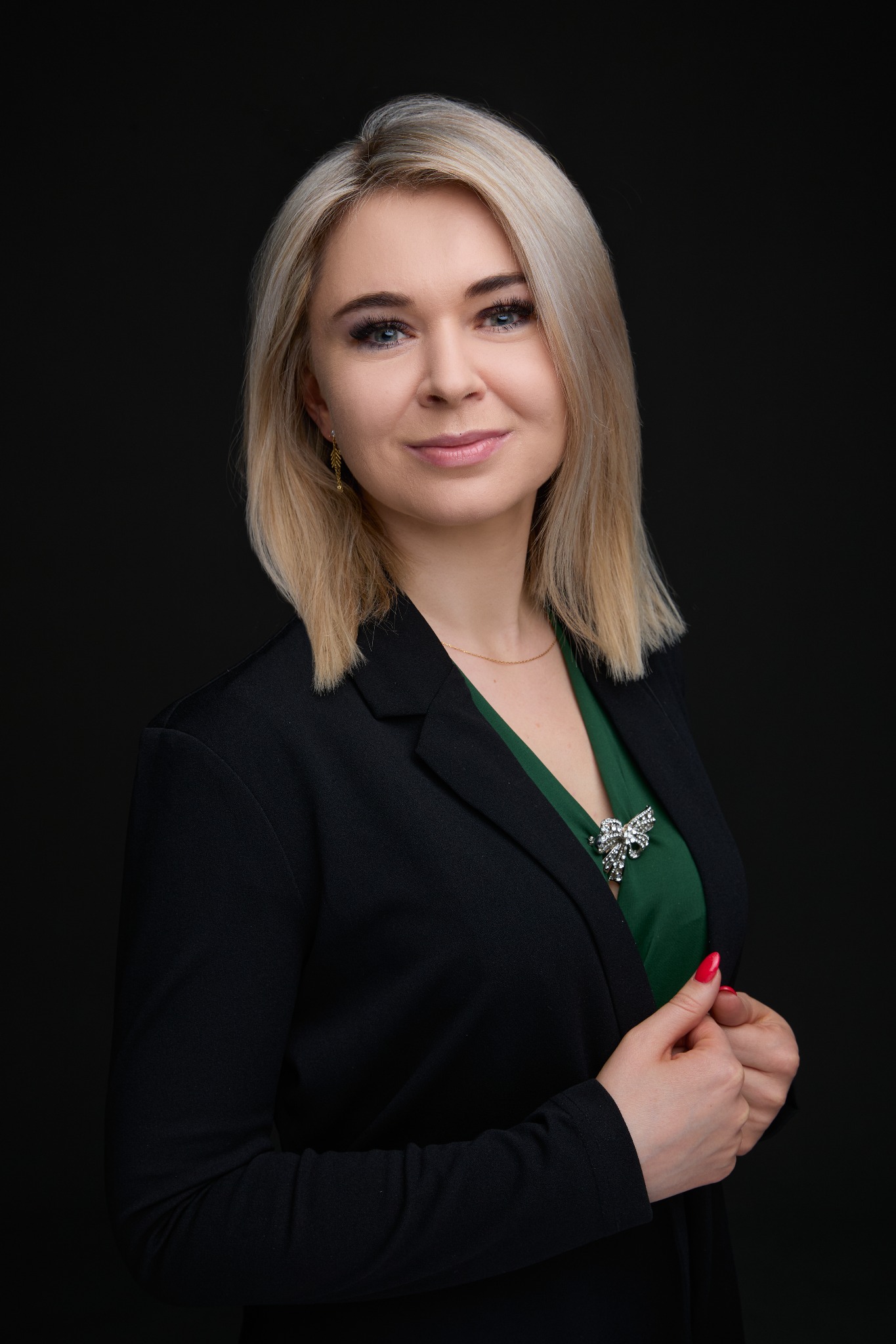 Portret uśmiechniętej blondwłosej kobiety w czarnej marynarce i zielonej bluzce z ozdobną broszką na ciemnym tle.