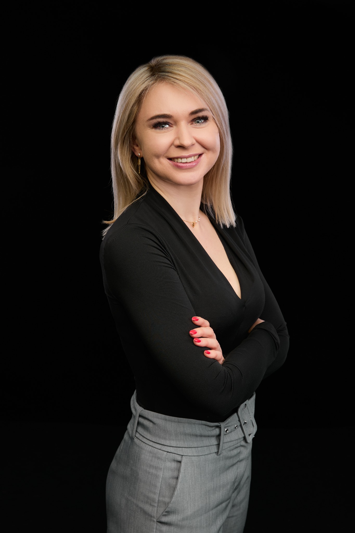 Portret uśmiechniętej blondynki w czarnej bluzce i szarych spodniach, z rękami skrzyżowanymi na piersiach na czarnym tle, z widocznym złotym łańcuszkiem.