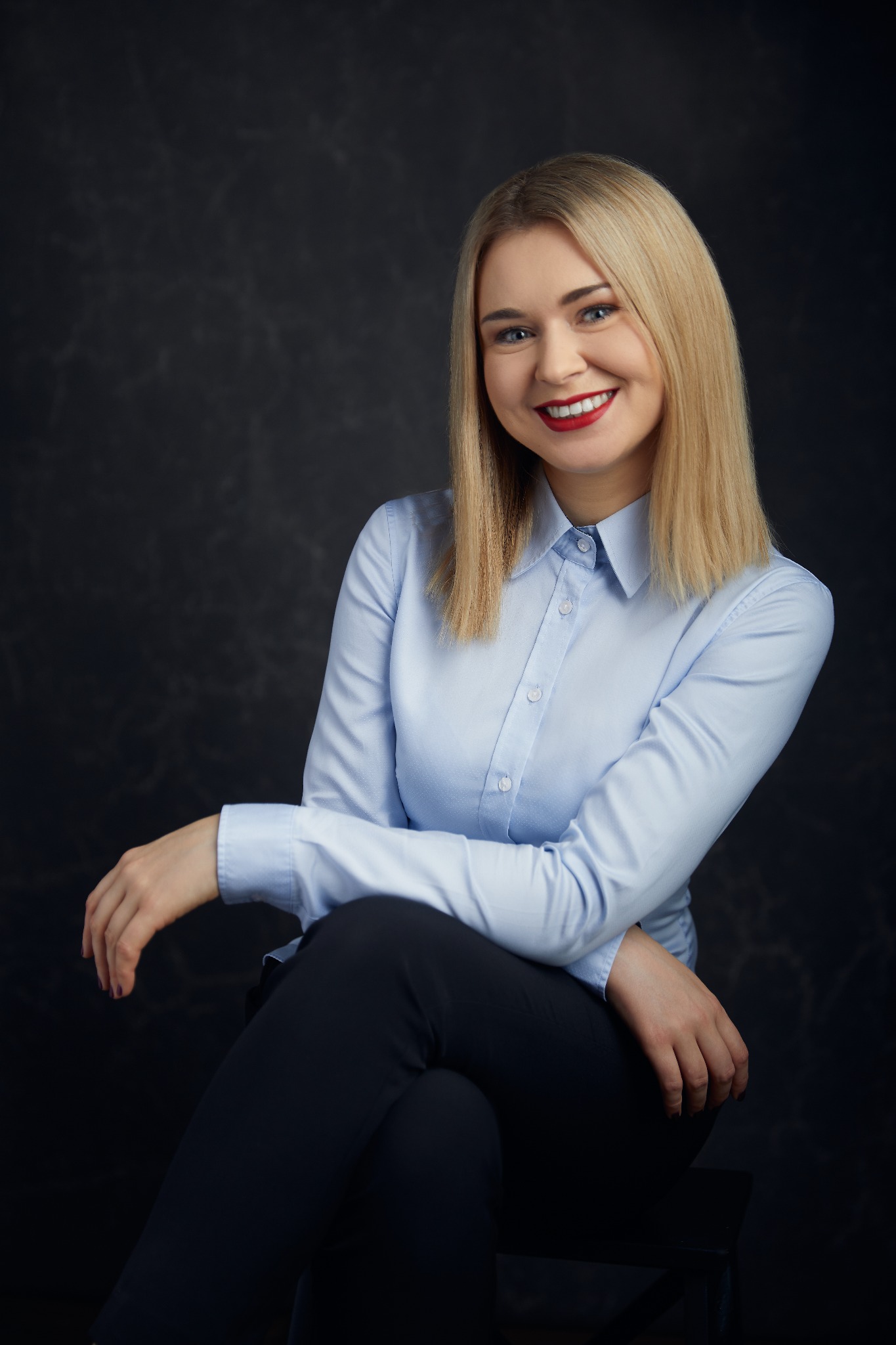 Portret uśmiechniętej kobiety w błękitnej koszuli i ciemnych spodniach, siedzącej na krześle na ciemnym tle.