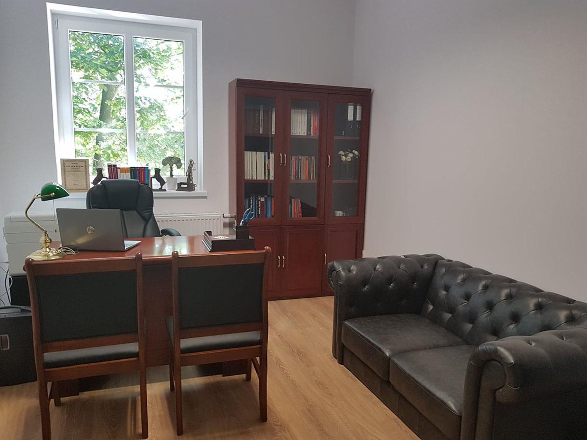 Gabinet z mahoniowym biurkiem, zieloną lampką, czarnym skórzanym fotelem, regałem z książkami i sofą typu chesterfield; widok przez okno na zieleń.