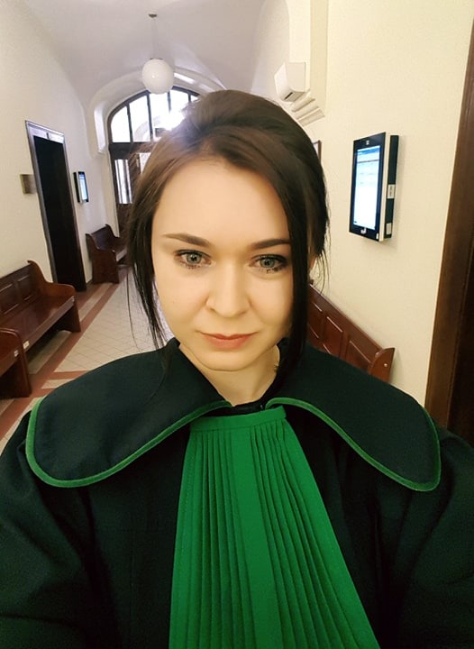 Portret kobiety w todze adwokackiej na tle korytarza z ławkami i urządzeniami elektronicznymi na ścianie.