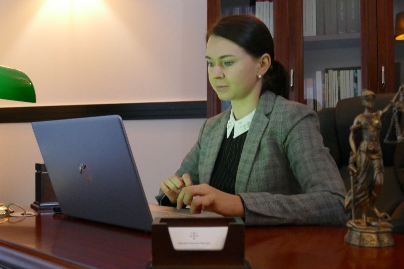 Kobieta w szarej marynarce, siedząca za biurkiem z laptopem, obok posążek Temidy i wizytownik z logo kancelarii.