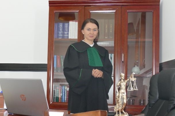 Kobieta w todze adwokackiej, stojąca w kancelarii na tle regału z książkami i figurki Temidy, z laptopem HP na biurku.