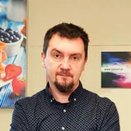 Portret mężczyzny z brodą, na drugim planie ściana z dwoma plakatami, jeden z motywem owoców, drugi z logo firmy lapisart.pl.