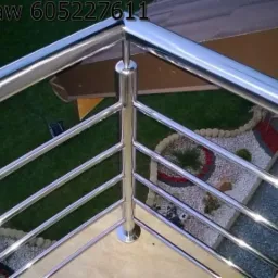 Błyszcząca, metalowa balustrada z poziomymi prętami, widok z góry na zadbany ogródek z kamykami i roślinami.