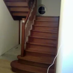 Drewniane schody z zaokrąglonym stopniem początkowym i balustradą z drewnianymi słupkami oraz metalowymi prętami, okrągłe okno na ścianie.