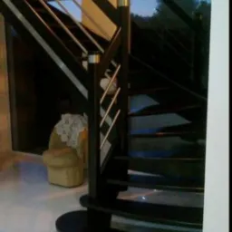 Ciemne, spiralne schody z drewnianymi stopniami i metalowymi poręczami w nowoczesnym wnętrzu, obok widoczny fotel z koronkową serwetą.