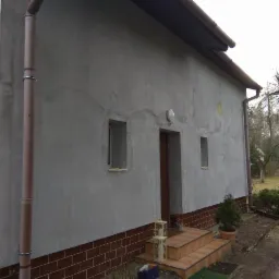 Budynek z szarą, surową elewacją, widoczne zacieki i nierówności, ceglana podmurówka, brązowe rynny, małe okna, lampa zewnętrzna, schody wejściowe z lampionami, trawnik i drzewa w tle.