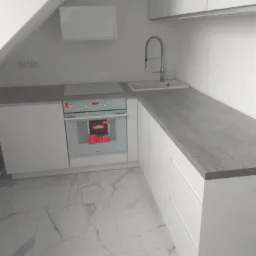 Biała kuchnia w nowoczesnym stylu, z marmurową podłogą i szarym blatem, umieszczona na poddaszu, widoczne białe szafki wiszące i piekarnik.