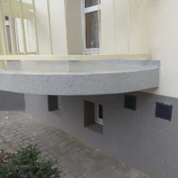 Zaokrąglony balkon z szarą, nakrapianą powierzchnią, wsparty na ścianie budynku z widocznymi małymi oknami i kratkami wentylacyjnymi, z kremową elewacją w górnej części.
