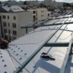 Montaż membrany PCV na dachu budynku wielorodzinnego z widocznymi elementami konstrukcji nośnej i narzędziami pracy. W tle zabudowa miejska.