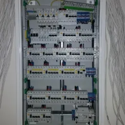 Rozdzielnica elektryczna w białej obudowie, widoczne liczne bezpieczniki, przewody i złączki, z wiązką kabli wchodzących z lewej strony, na jasnej ścianie.