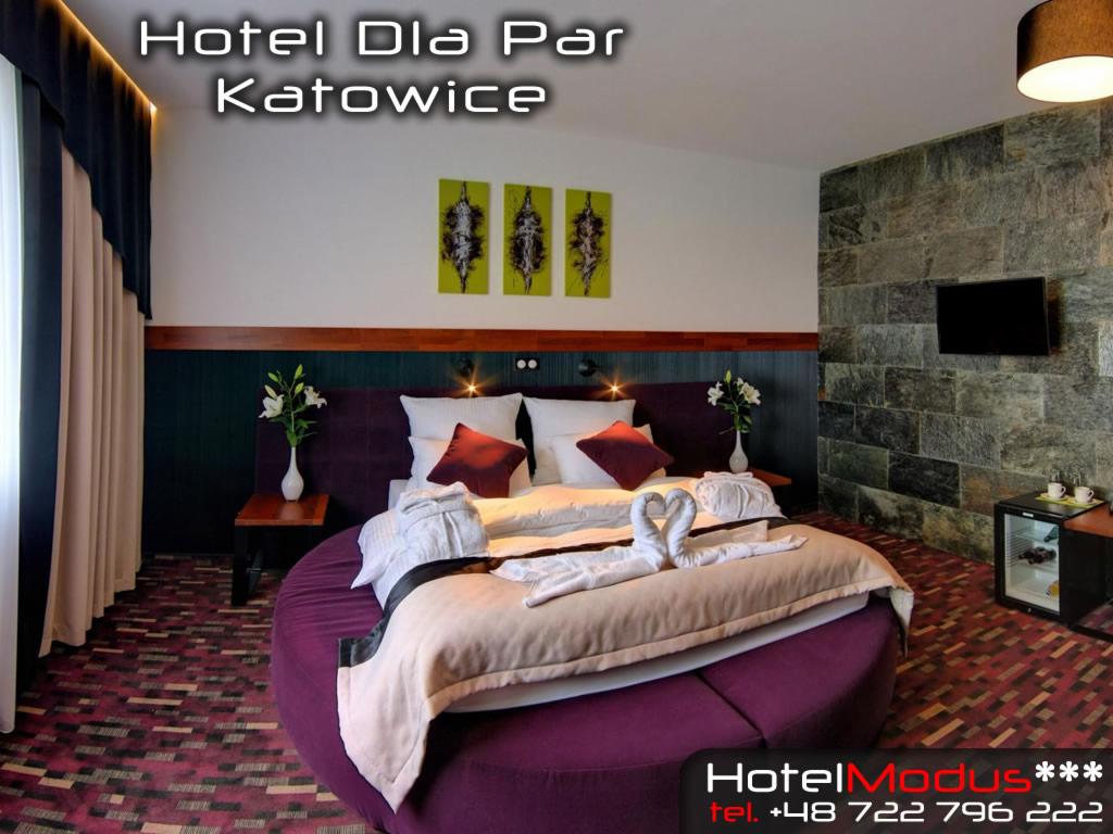 Pokój hotelowy z okrągłym łóżkiem w kolorze fioletowym, ręczniki ułożone w kształt łabędzi, ściana z kamienia i mała lodówka.