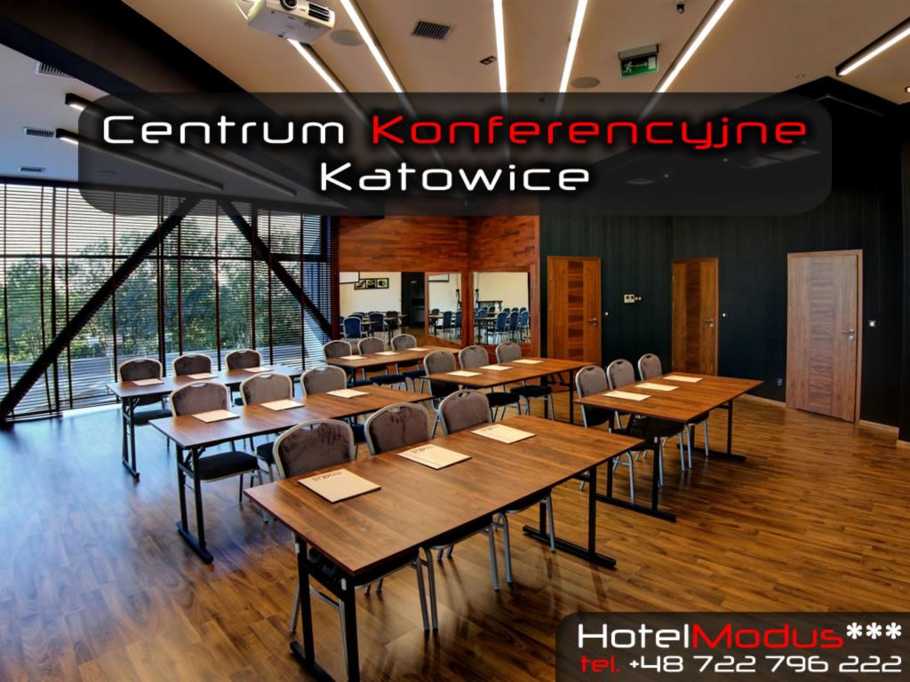 Sala konferencyjna z drewnianymi stołami i krzesłami, oświetlona lampami sufitowymi, z widokiem na zewnątrz przez okna z żaluzjami, w tle drzwi i lustro.