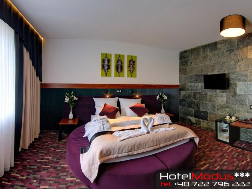 Pokój hotelowy z okrągłym łóżkiem, pościel ułożona w kształt łabędzi, ściana z kamienia i minimalistyczne obrazy.