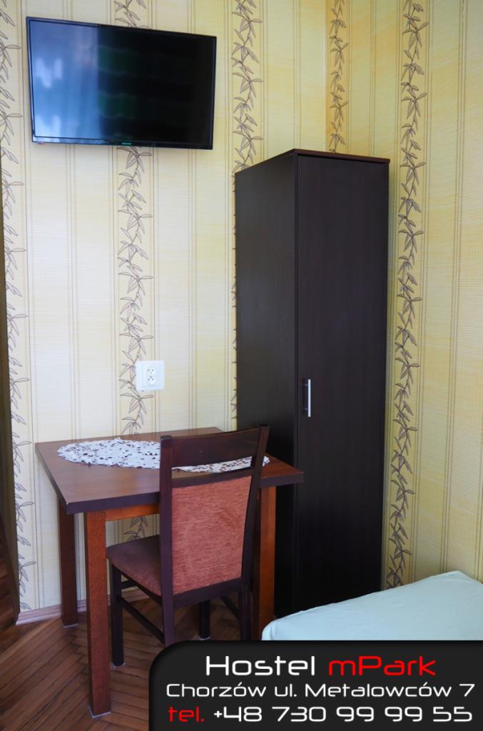 Wnętrze pokoju w hostelu: biurko z serwetką, krzesło, szafa i telewizor na ścianie z wzorzystą tapetą.
