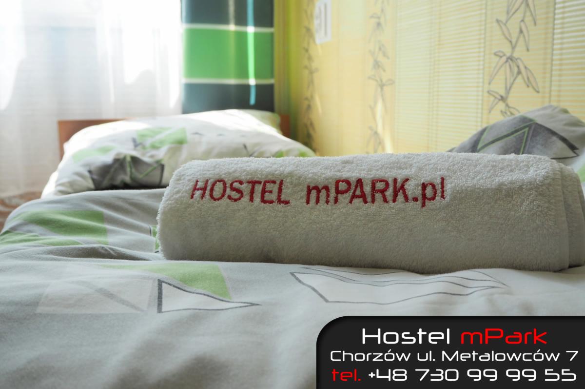 Pokój w hostelu z ręcznikiem z wyhaftowaną nazwą i adresem 'HOSTEL mPARK.pl' leżącym na łóżku z pościelą w geometryczne wzory.