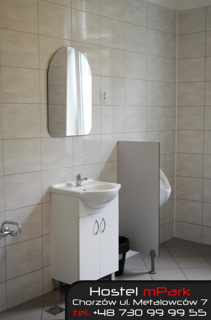 Wnętrze łazienki w hostelu: umywalka z szafką, owalne lustro na ścianie z jasnych płytek, pisuar za szarą ścianką działową.