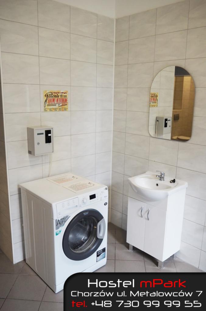 Wnętrze łazienki w hostelu z białą pralką, umywalką z szafką, lustrem i plakatem na kafelkowej ścianie.