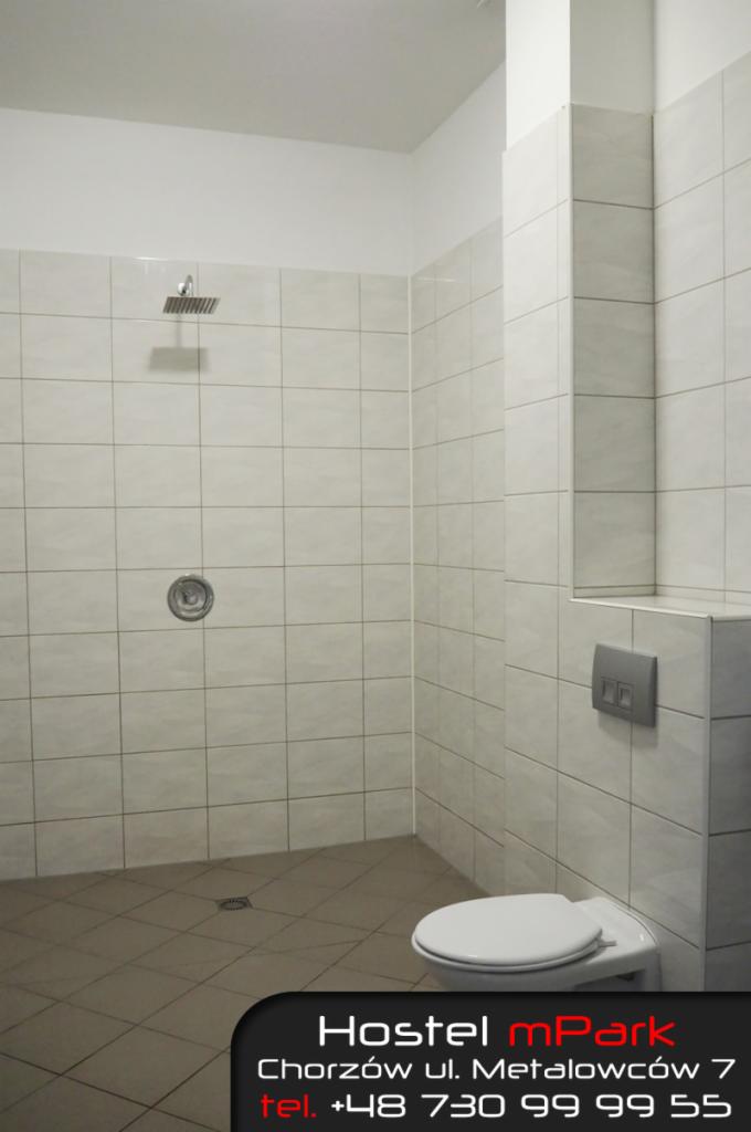 Wnętrze łazienki w hostelu z kabiną prysznicową i toaletą, wykończonej białymi i brązowymi płytkami, z widocznym logo i adresem hostelu w dolnej części zdjęcia.