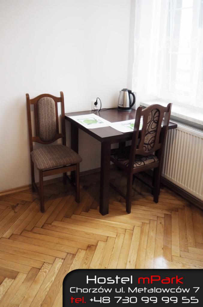 Pokój w hostelu: drewniany stół, dwa krzesła z tapicerowanymi siedziskami i oparciami, czajnik elektryczny na blacie, parkiet w jodełkę na podłodze.