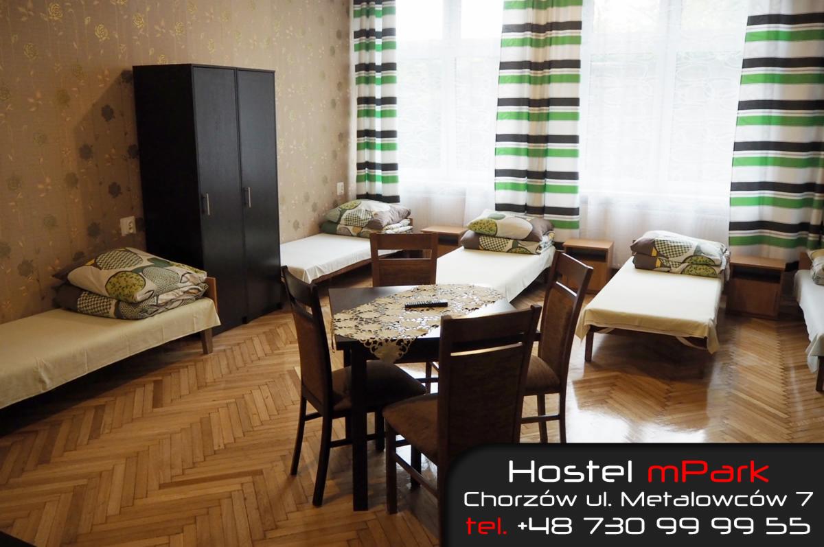 Wnętrze pokoju w hostelu: cztery łóżka pojedyncze z pościelą, szafa, stół z czterema krzesłami, okno z zasłonami w zielono-białe pasy, parkiet na podłodze, tapeta w kwiaty.