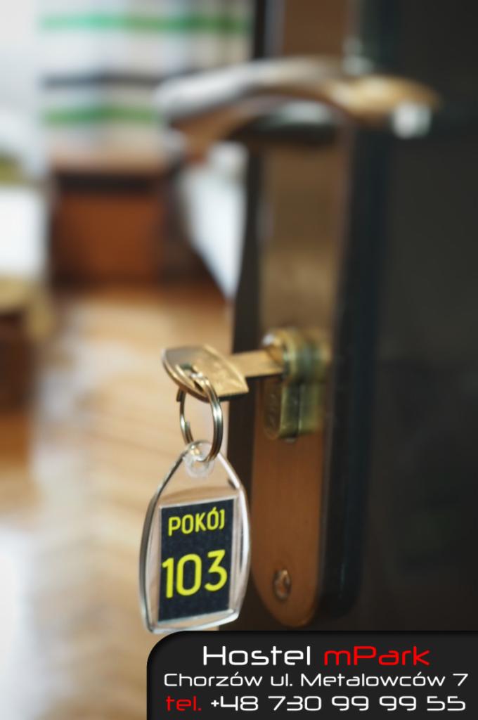 Klucz z brelokiem z numerem pokoju 103 w zamku drzwi hostelu mPark, Chorzów. Widoczny numer telefonu.