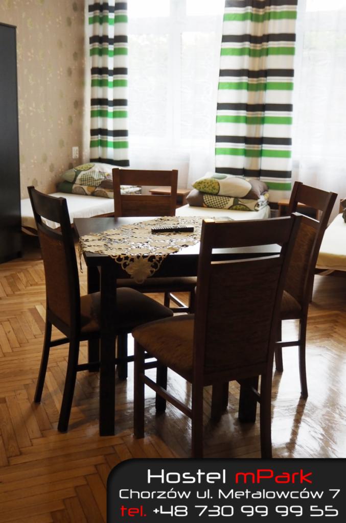 Wnętrze pokoju w hostelu: stół z koronkową serwetą, cztery krzesła, łóżko z poduszkami w tle, okno z zasłonami w paski, parkiet na podłodze. W dolnej części kadru logo hostelu z adresem i numerem...