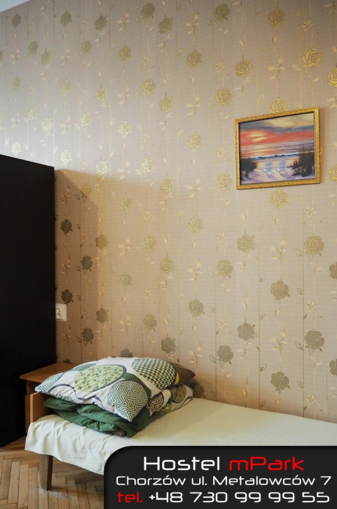 Pokój w hostelu z łóżkiem, poduszkami w zielone wzory i obrazkiem nad łóżkiem przedstawiającym plażę o zachodzie słońca, ściany pokryte tapetą w róże.