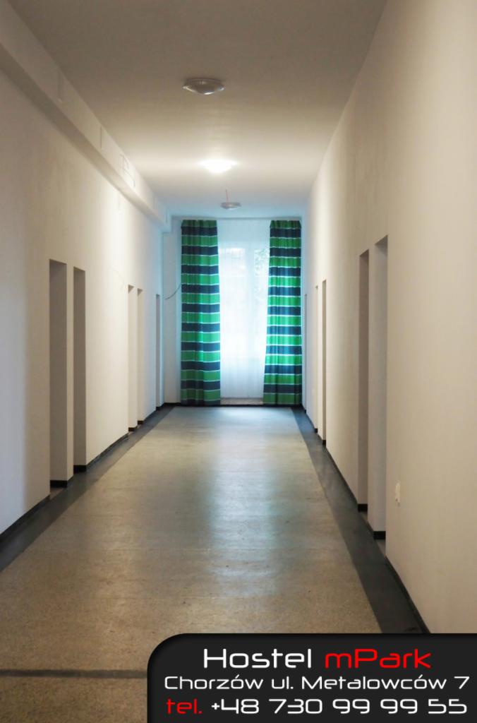 Wnętrze długiego, pustego korytarza w hostelu z drzwiami po obu stronach i oknem z zielono-niebieskimi zasłonami na końcu. Na dole kadru widoczny fragment logo i danych kontaktowych hostelu.
