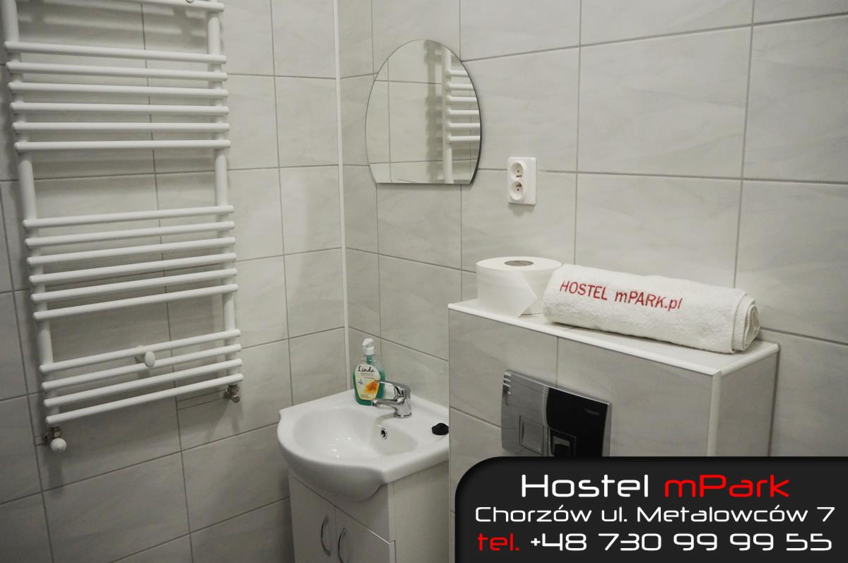 Łazienka w hostelu: białe płytki, umywalka z szafką, grzejnik drabinkowy, okrągłe lustro, ręcznik z logo 'HOSTEL mPARK.pl'.