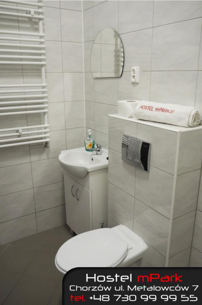 Jasna łazienka w hostelu z umywalką, toaletą, lustrem i grzejnikiem drabinkowym, ręcznik z logo hostelu na górze.