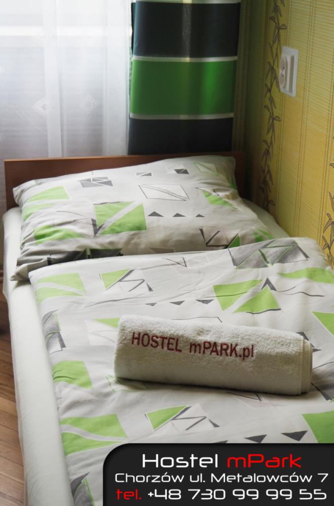 Pokój w hostelu z pojedynczym łóżkiem, pościel w geometryczne wzory w odcieniach zieleni i szarości, na łóżku zwinięty biały ręcznik z wyhaftowanym napisem 'HOSTEL mPARK.pl', w tle okno z firanką...