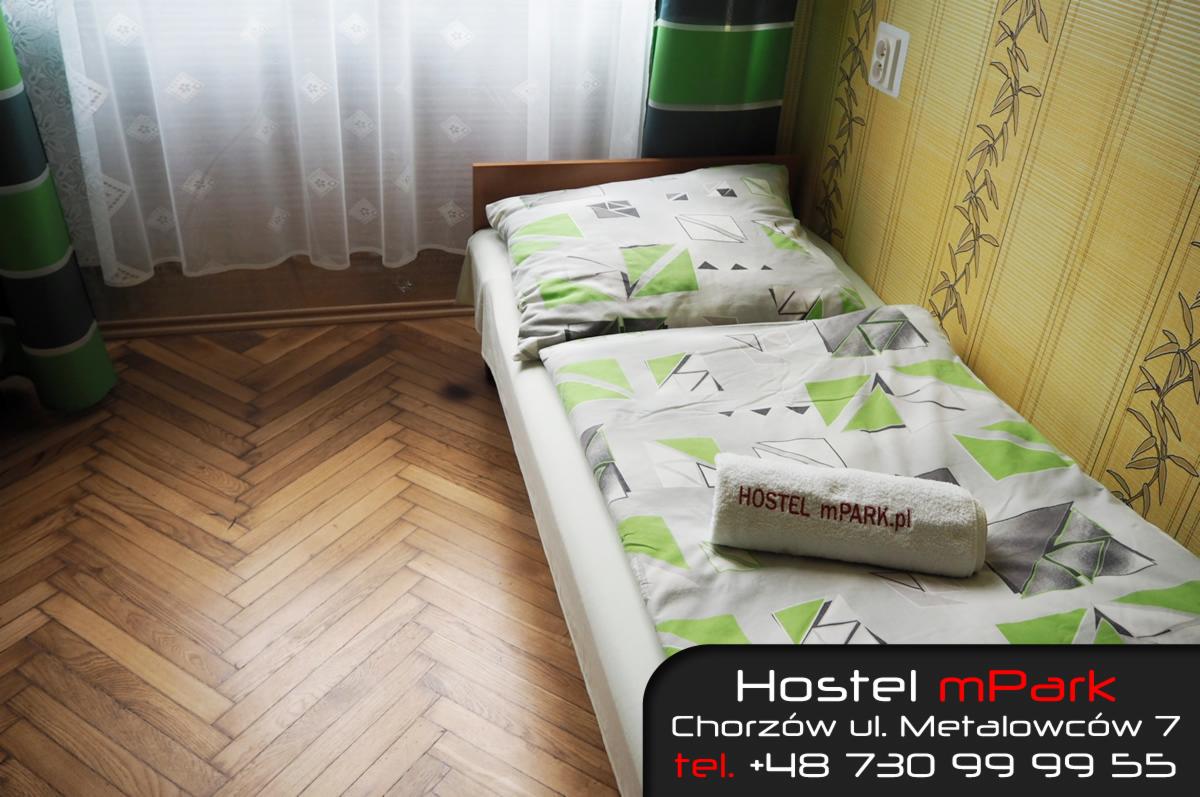 Pokój hostelowy z pojedynczym łóżkiem, pościel w geometryczne wzory zieleni i szarości, ręcznik z logo hostelu, parkiet w jodełkę, zasłony w tle.