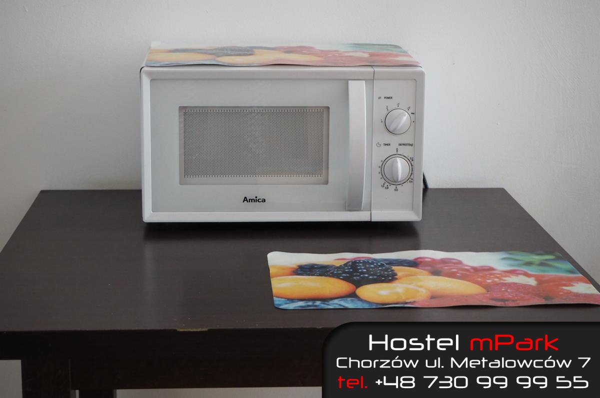 Biała kuchenka mikrofalowa marki Amica na ciemnym stole, z podkładką z motywem owoców pod spodem i na górze, widoczny napis 'Hostel mPark' z adresem i numerem telefonu.
